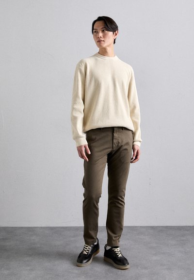 Pull en maille côtelée, pantalon vert olive, baskets noires avec semelles claires. Pose debout, fond neutre, texture lisse, design décontracté.