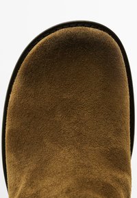 Stivale in suede marrone con punta arrotondata, superficie testurizzata e suola in gomma nera. I dettagli delle cuciture lungo il bordo aggiungono interesse visivo.