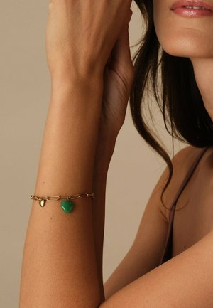 Kvinde iført et guldarmbånd med et grønt hjertevedhæng på sin hævede underarm, hvor en del af hendes ansigt og skulder ses.