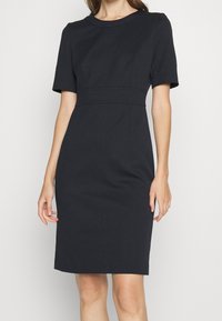 Robe ajustée bleu marine foncé avec manches courtes, encolure ronde et coutures décoratives. Longueur genou avec une silhouette structurée et tissu texturé.