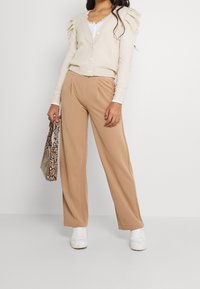 Kobieta w beżowym kardiganie i szerokich spodniacach w kolorze camel, trzymająca torebkę w wężowy wzór, stojąca w białych sneakersach na jednolitym tle.