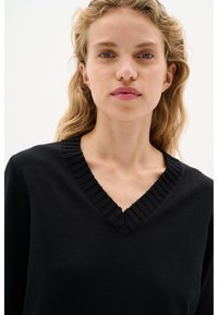 Donna bionda con capelli mossi che indossa un maglione nero a V, rivolta di fronte con un'espressione neutra su uno sfondo bianco uniforme.