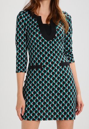 Frau trägt ein enganliegendes schwarzes Kleid mit blau-weißem geometrischem Muster, dreiviertel Ärmeln und schwarzer Einfassung am Halsausschnitt und den Taschen.