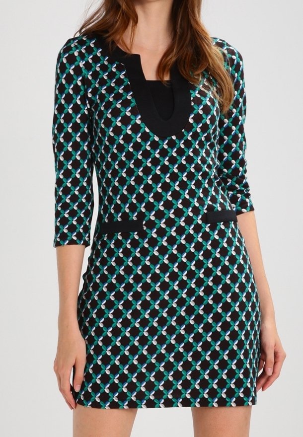 Femme portant une robe noire cintrée avec un motif géométrique bleu et blanc, manches trois-quarts et bordure noire autour de l'encolure et des poches.