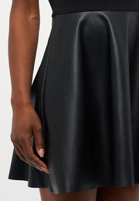 Detailní záběr na tmavou ruku odpočívající vedle černé kožené sukně s jemnými záhyby a hladkou texturou.