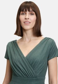 Zielona sukienka z krótkimi rękawami, głębokim dekoltem w serek oraz marszczeniami w okolicy biustu, wykonana z teksturowanej, połyskującej tkaniny.