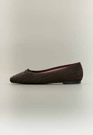Ballerine plate en cuir tressé marron foncé avec un petit nœud sur le bout arrondi et un talon bas et plat sur un fond neutre.