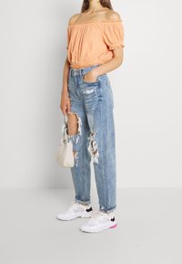 Top arancione strutturato, spalline scoperte; jeans a vita alta blu chiaro con grandi strappi; sneakers bianche con accenti rosa; borsa piccola beige.