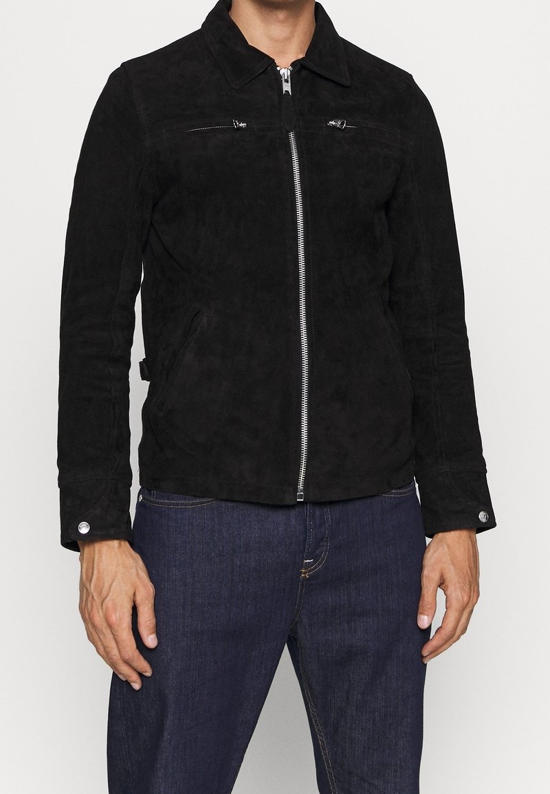 Homme portant une veste zippée en suède noir avec col et un jean bleu, debout devant un fond clair uni.