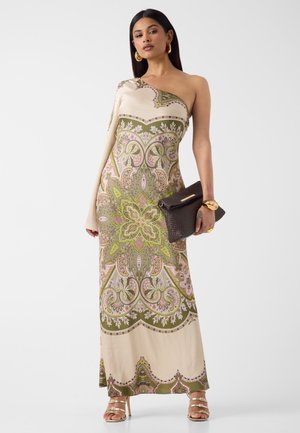 Femme portant une robe longue asymétrique avec un motif orné vert et rose, tenant une pochette texturée foncée, accessoirisée avec des bijoux en or et des talons.