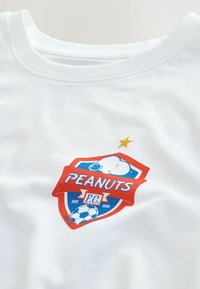 T-shirt blanc avec un logo Peanuts F.C. rouge et bleu représentant Snoopy, un ballon de football et une étoile dorée au-dessus de l'emblème.