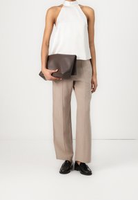 Haut halter blanc, pantalons larges beiges et mocassins noirs. Tenant une pochette en cuir marron avec une forme légèrement arrondie et des détails embossés.