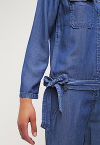 Džínový overal středně modré barvy s dlouhými rukávy, předními kapsami a detaily na zavazování v pase. Materiál je měkký na omak s klasickým vzhledem.