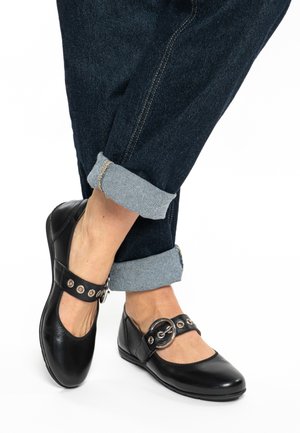 Schwarze flache Lederschuhe mit rundlicher Spitze, einem Riemen mit silberner Beschlagsverzierung und offenen Seiten. Kombiniert mit hochgekrempelten Jeanshosen.