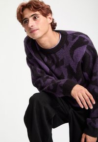 Suéter de cuello redondo en tonos morados y negros con un patrón, hecho de un material texturizado, acompañado de pantalones negros. La figura está posada de rodillas.