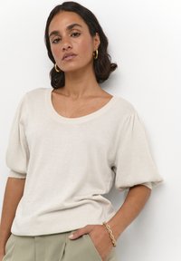 Helles beige gestricktes Sweatshirt mit Rundhalsausschnitt, kurzen Puffärmeln und einer weichen Textur. Getragen mit gefalteten olivgrünen Hosen und einem Armband.