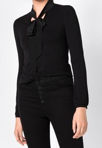 Svart långärmad topp med design av slipsknut vid halsen; mjukt, stretchigt tyg med en figurnära silhuett. Kombinerad med högmidjade svarta jeans med knapppdetail.