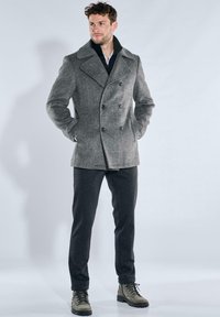 Grauer Woll-Peacoat mit zweireihigem Design, weitem Kragen und schwarzen Knöpfen, kombiniert mit dunkler Hose und olivgrünen Stiefeln.