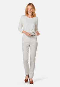 Witte long-sleeve top gecombineerd met lichtbeige broeken en bruin suède hakken, staande met een bril in de hand, glimlachend tegen een effen achtergrond.