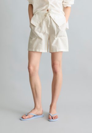 Persoon draagt een off-white overhemd met knopen en shorts met trekkoord, gecombineerd met lichtblauwe slippers, staand op een grijze achtergrond.