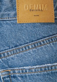 Blue denim fabric with orange stitching and a tan leather label embossed with "DENIM" and printed with "Bershka Est. 1998".