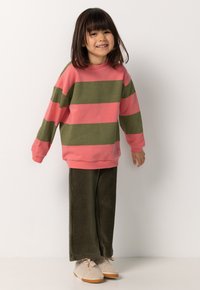 Ein lächelndes Kind mit schulterlangem Haar trägt einen pink- und grün gestreiften Pullover, grüne Cordhosen und beige Sneakers vor einem weißen Hintergrund.