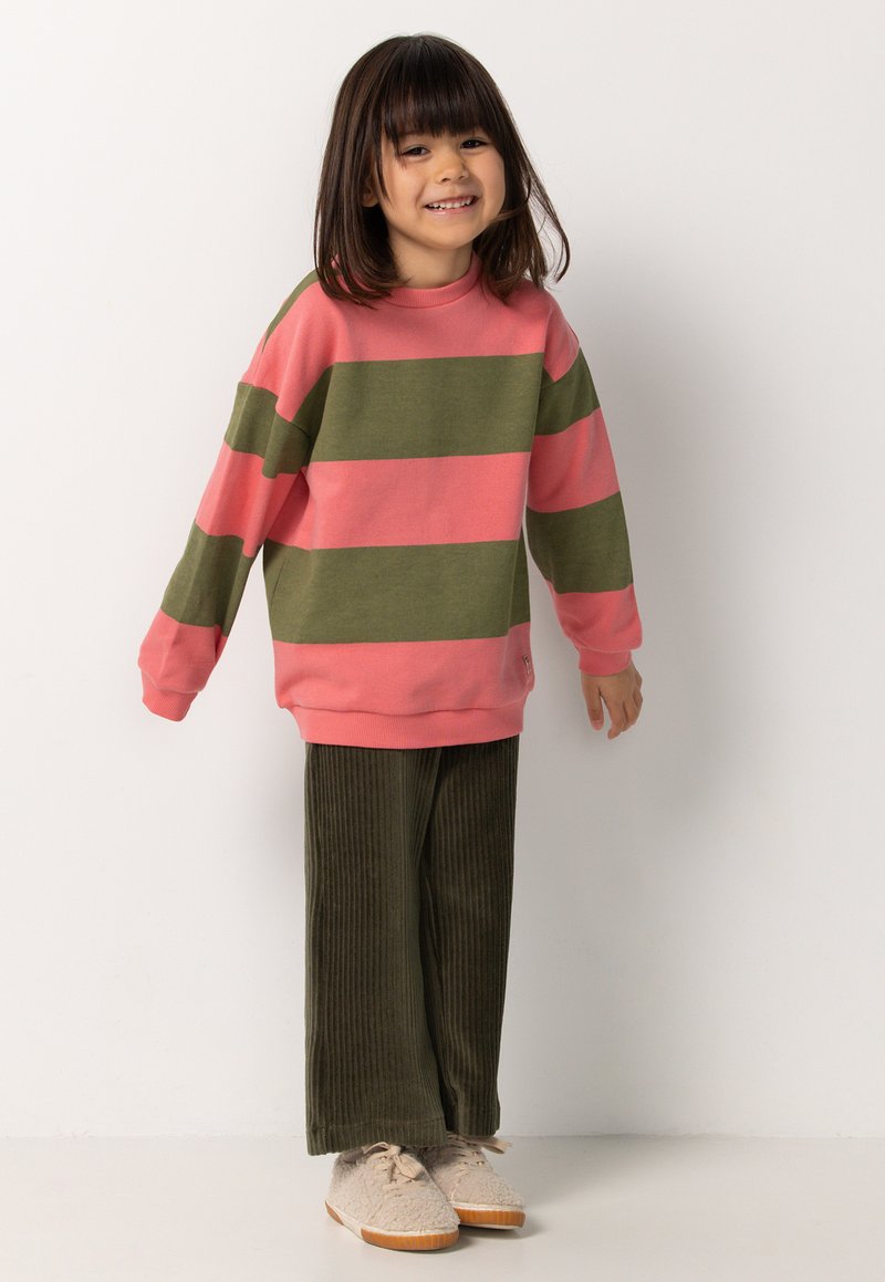 Ein lächelndes Kind mit schulterlangem Haar trägt einen pink- und grün gestreiften Pullover, grüne Cordhosen und beige Sneakers vor einem weißen Hintergrund.