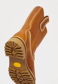 Ruskea nahkasaapas, jossa on kestävä kumipohja ja yksityiskohtainen tikkaus. Kuva on vinosti otettu, jolloin näkyvät teksturoidut kuminastat ja vetolenkki varren yläosassa.