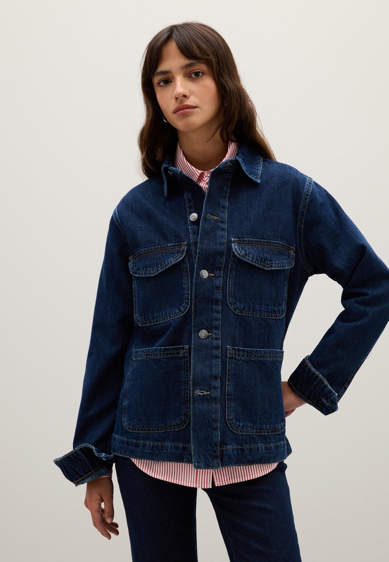 Donna con lunghi capelli castani che indossa una giacca di jeans blu scuro sopra una maglietta a righe rossa e bianca, in piedi con una mano sull'anca.