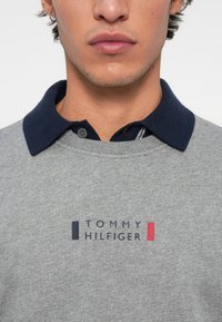 Lähivõte inimesest, kes kannab halli dressipluusi, millel on "Tommy Hilfiger" logo, ja selle all on tume sinine kraega särk.