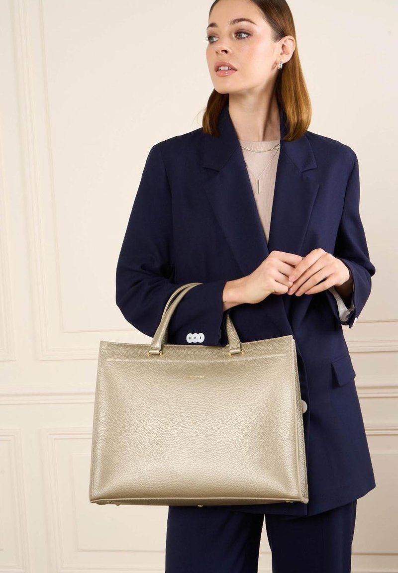Femme en costume bleu marine tenant un grand sac à main en cuir beige, regardant sur le côté devant un mur clair avec des panneaux.