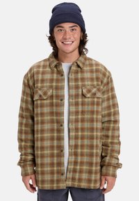 Braun-gelb kariertes Flanellhemd mit zwei Brusttaschen, Knopfleiste und langen Ärmeln, getragen über einem hellgrauen T-Shirt.