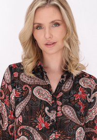 Blusa nera con motivo paisley in rosso, bianco e grigio. Caratterizzata da bottoni e un colletto. Tessuto leggero con un leggero lucido.
