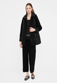 Cappotto trapuntato nero con cappuccio e chiusura a zip, abbinato a un top nero con una striscia bianca, pantaloni neri larghi e scarpe nere.
