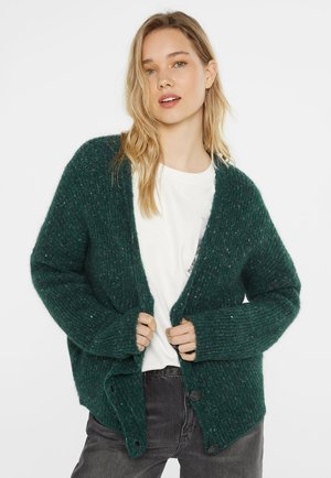 Young woman with blonde hair wearing a dark green knitted cardigan over a white shirt and dark gray pants, standing against a plain white background.