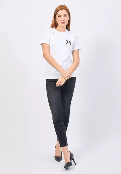 T-shirt bianco a maniche corte con grafica nera a forma di 'X' e dettagli dorati, abbinato a jeans neri slim-fit e décolleté nere a punta.