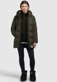 Gepolsterte olivgrüne Parka mit Kapuze, ausgestattet mit einem Reißverschluss vorne, schwarzen Akzenten und einem Logo-Patch. Kombiniert mit einem dunklen Pullover und schwarzen Leggings.