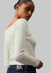 Top de manga larga canalé en color blanco roto, con un escote amplio y una silueta ajustada, combinado con unos vaqueros de mezclilla azul oscuro que presentan un parche con el logo.