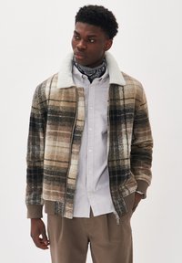 Jachetă din material tartan maro și bej, cu guler din fleece, închidere cu fermoar și manșete cu nervuri. Purtați deasupra unei cămăși albe cu dungi și pantaloni bej.
