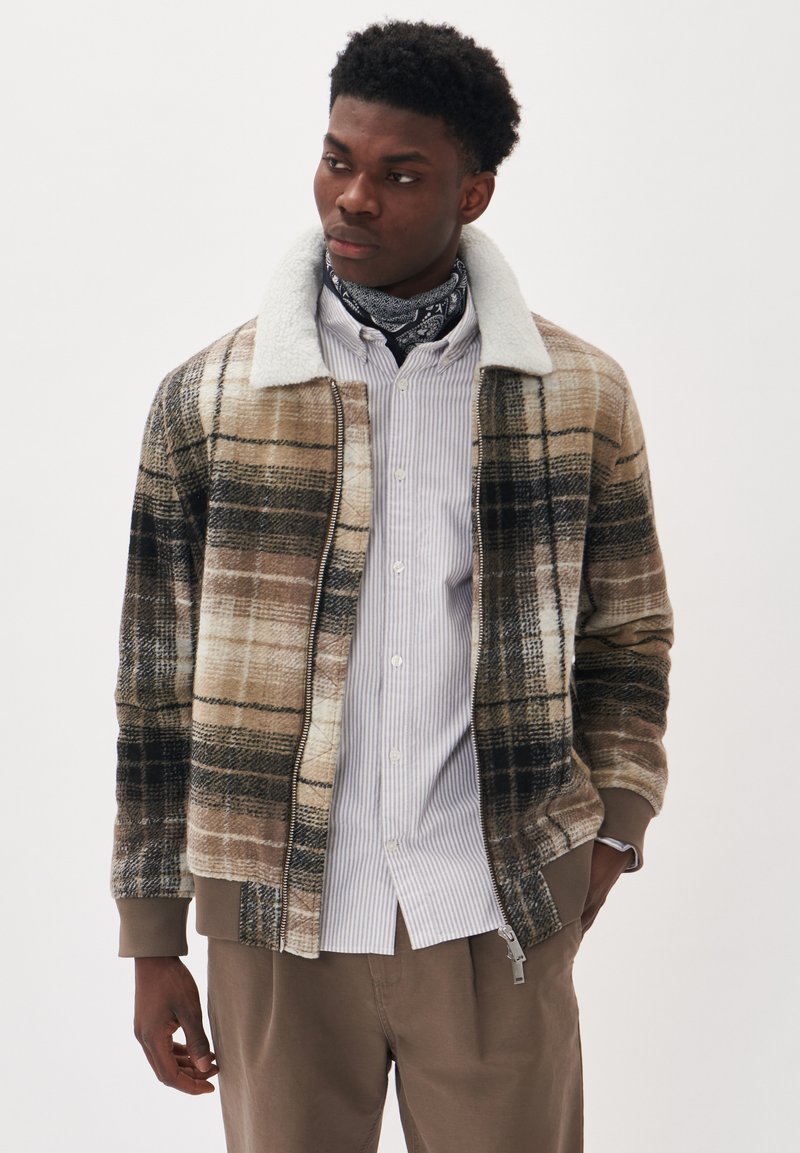 Jachetă din material tartan maro și bej, cu guler din fleece, închidere cu fermoar și manșete cu nervuri. Purtați deasupra unei cămăși albe cu dungi și pantaloni bej.