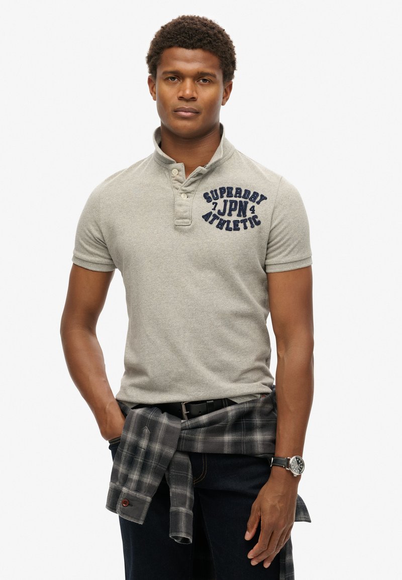 Polo shirt gris à manches courtes avec col, arborant le texte brodé bleu marine "SUPERDRY JPN ATHLETIC" sur la poitrine gauche. Porté avec une ceinture noire.