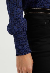 Manžeta rukávu modré blůzy s dlouhým rukávem, s nařasenou elastickou částí a černými nepravidelnými skvrnami, kombinovaná s černými kalhotami a tenkým černým páskem se zlatým lemováním.