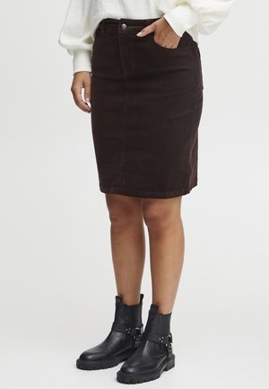 Vrouw draagt een donkerbruine rok tot op de knie, een wit shirt met lange mouwen en zwarte enkellaarsjes met gespen, staand tegen een witte achtergrond.