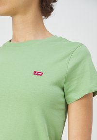 Personne portant une chemise à manches courtes vert clair avec une petite étiquette rouge du logo Levi's sur la poitrine, tournée vers la gauche.