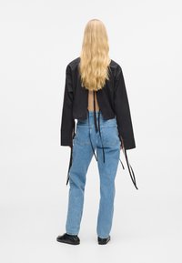 Čierny kratký top s otvoreným chrbtom a viazaním, v kombinácii so svetlomodrými džínsami. Modelka má dlhé vlnité vlasy, styled dozadu.