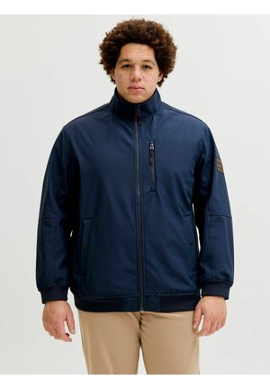 Młody mężczyzna z kręconymi włosami, ubrany w granatową kurtkę na zamek i beżowe spodnie, stojący na jednolitym jasnym tle.