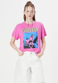 Rosa bomulls t-skjorte med grafisk trykk av et band i blått og svart. Korte ermer, avslappet passform, sammen med hvite denim shorts.