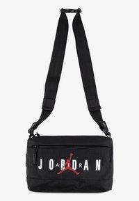 Sac en toile noir à bandoulière avec sangle ajustable, poche zippée sur le devant, texte "JORDAN" en blanc audacieux et détail du logo "Jumpman" rouge.