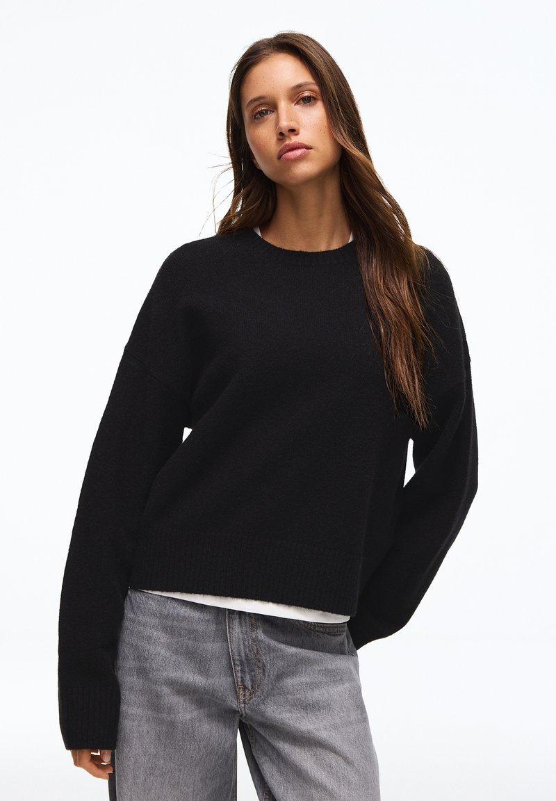 Jeune femme aux longs cheveux bruns portant un pull noir et un jean gris délavé, debout devant un fond blanc.