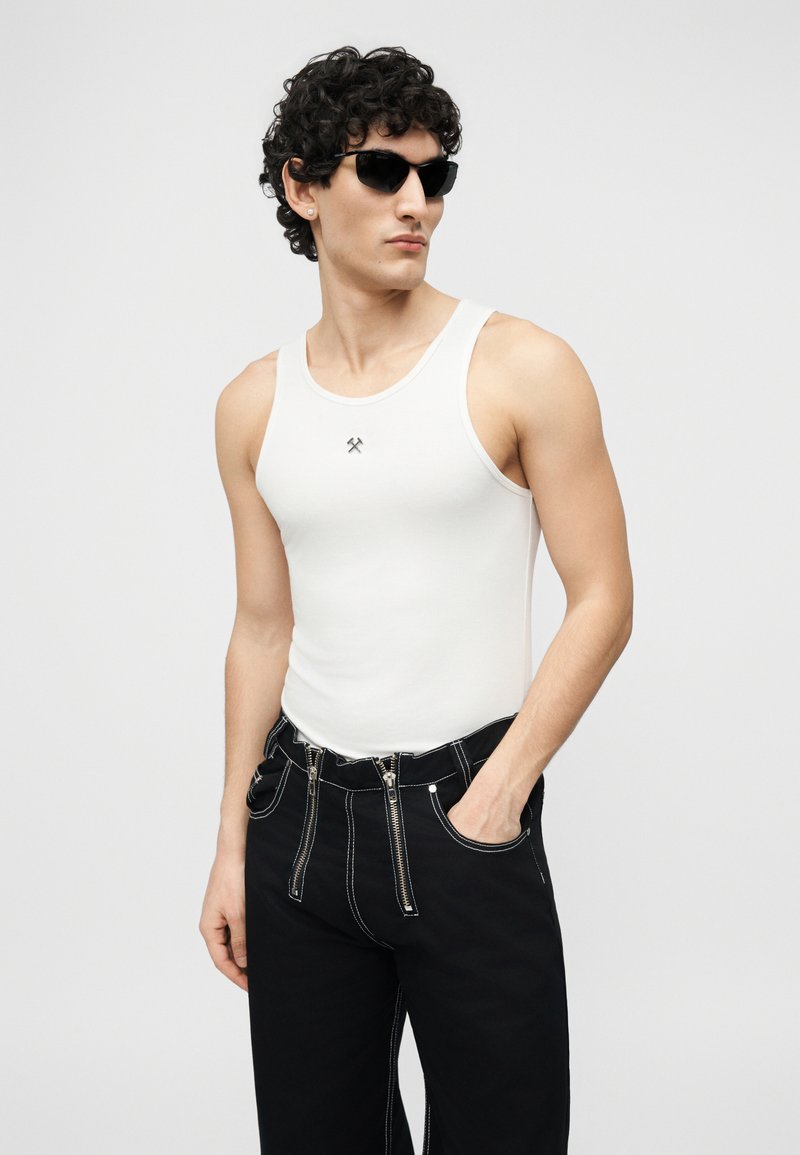 Man wearing black sunglasses, white tank top with small logo, and black pants with exposed silver zippers, standing with one hand in pocket.
